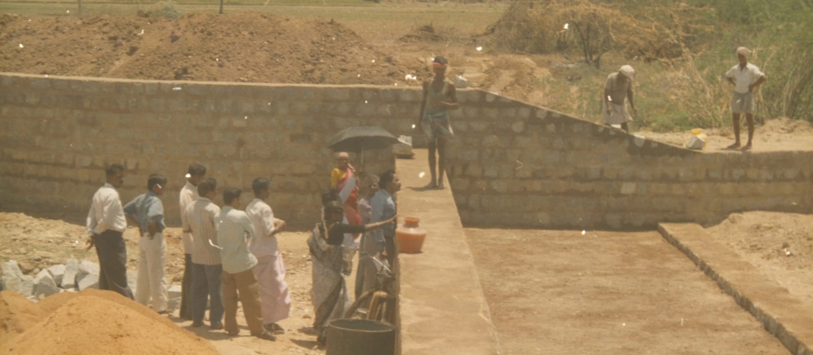 A masonry water-harvesting structure under construction, its block walls part-built, with heaps of sand and aggregate in front and two labourers working on the wall