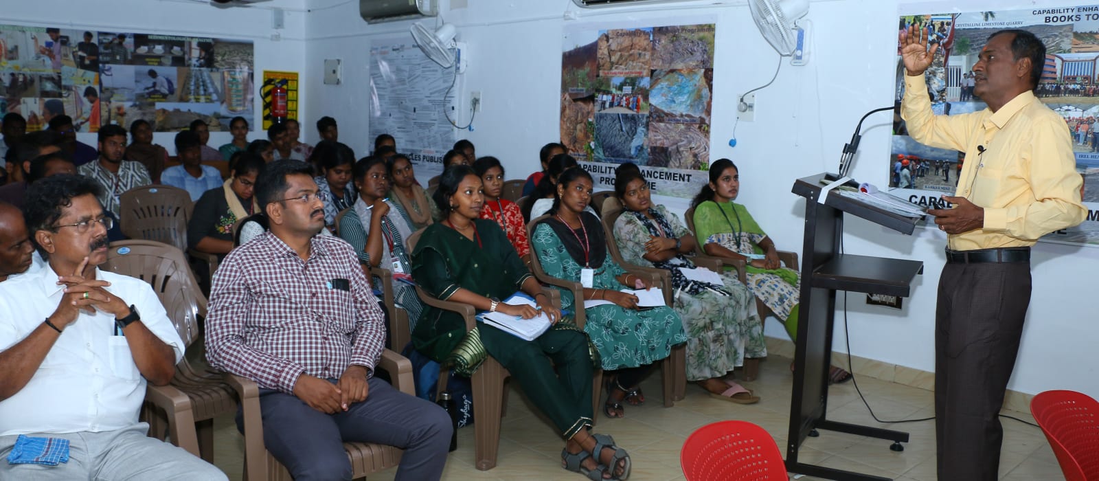 A trainer at a standing lectern with one hand raised mid-point, addressing rows of seated women participants who are writing in notebooks, with senior officials listening at the left