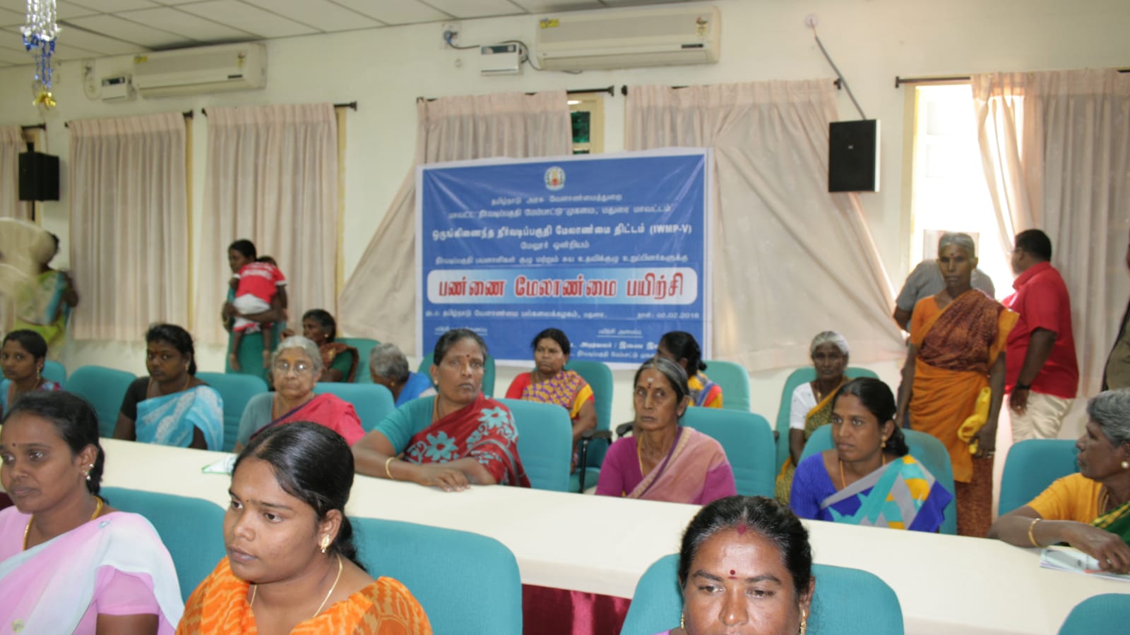 Rows of women trainees seated at tables in an auditorium, several faces in the front rows sharp and attentive