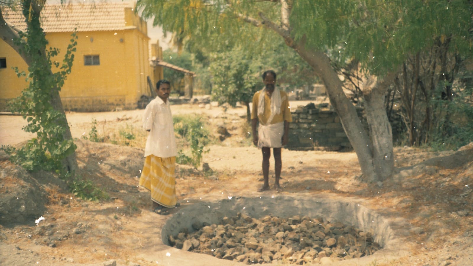 A stone-filled percolation pit dug beside a village lane under tamarind trees, with two men in dhotis standing on its rim and a yellow-washed village house behind