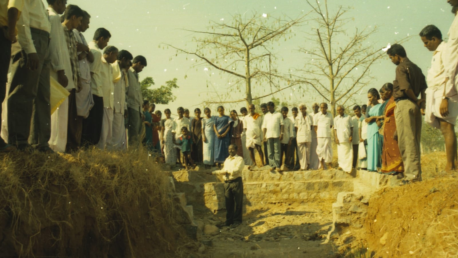 About fifty villagers, men and women, standing along both banks at the top of a freshly cut waterway while one man stands down in the excavated channel beside a stone check structure