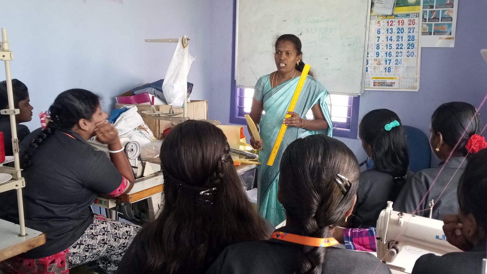 A woman instructor at a whiteboard holding a measuring tape, teaching a garment-making class
