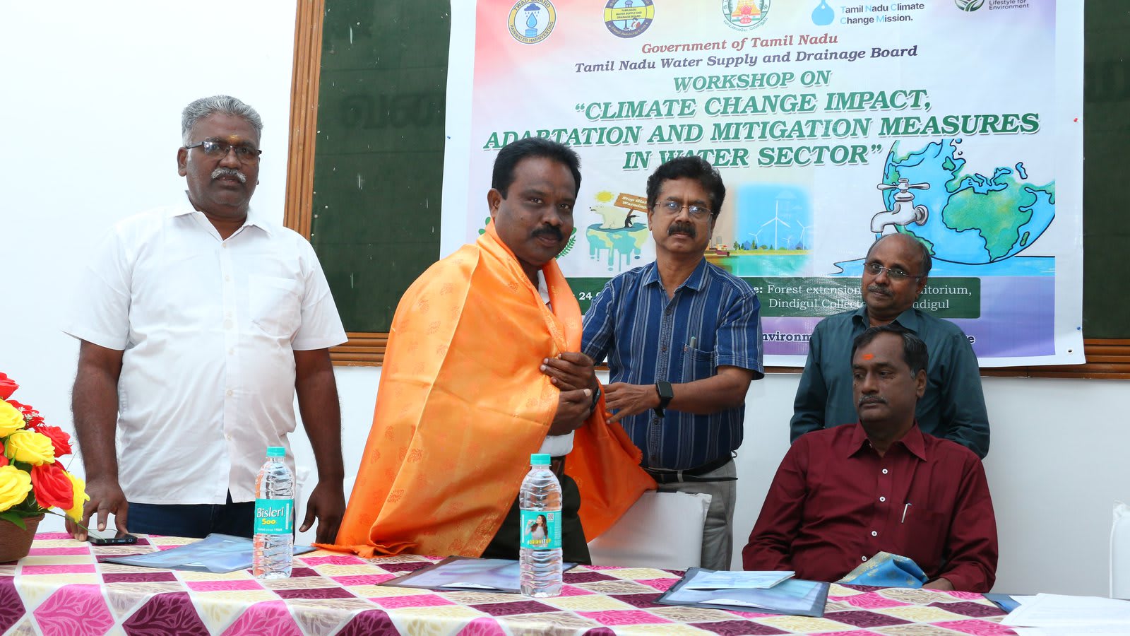 Four men at a dais, one of them draped in an orange shawl, in front of a printed banner reading &ldquo;Government of Tamil Nadu, Tamil Nadu Water Supply and Drainage Board &mdash; Workshop on &lsquo;Climate Change Impact, Adaptation and Mitigation Measures in Water Sector&rsquo;&rdquo;