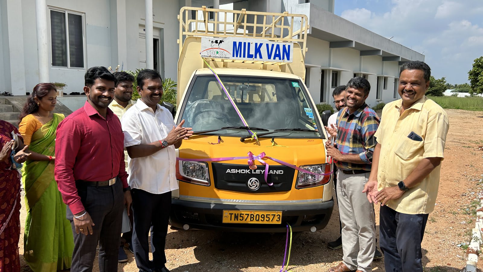 A yellow Ashok Leyland van lettered &ldquo;Milk Van&rdquo; and tied with a purple ribbon, with men and women standing on either side of it outside a long single-storey building