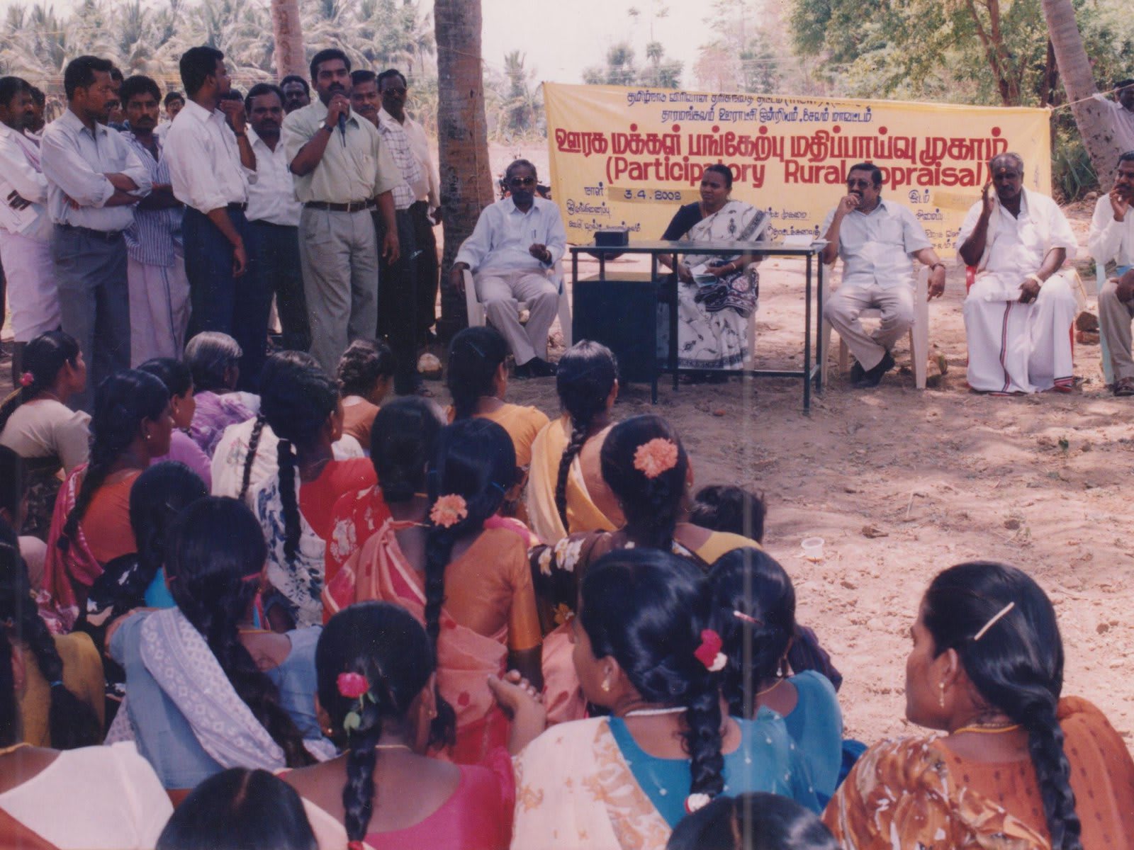 A large group of women and girls seated on the ground outdoors facing a table under a cloth banner, men standing behind