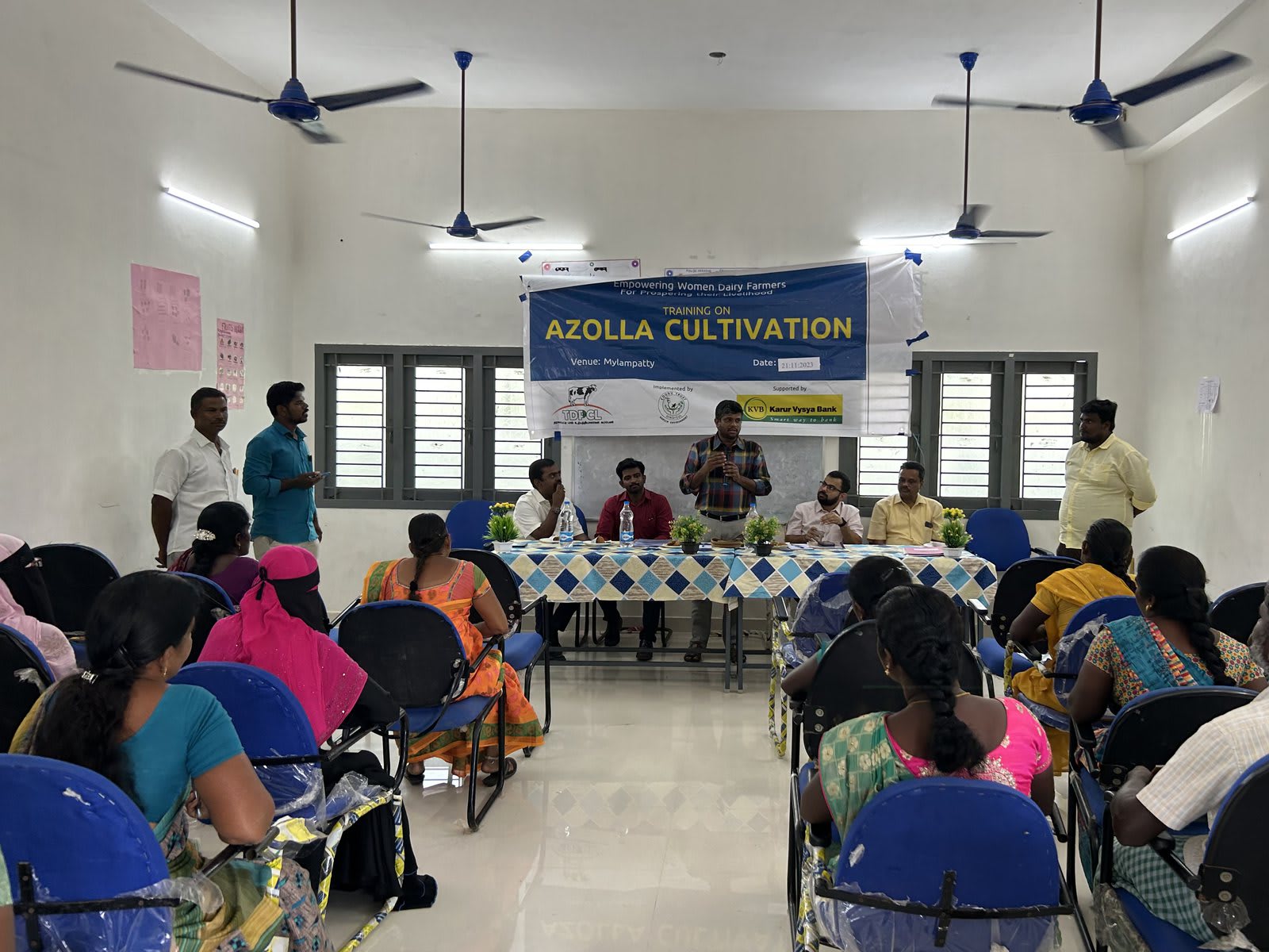 Rows of women seated on plastic chairs in a bright hall facing a table of speakers, with a printed banner reading Azolla Cultivation on the wall behind
