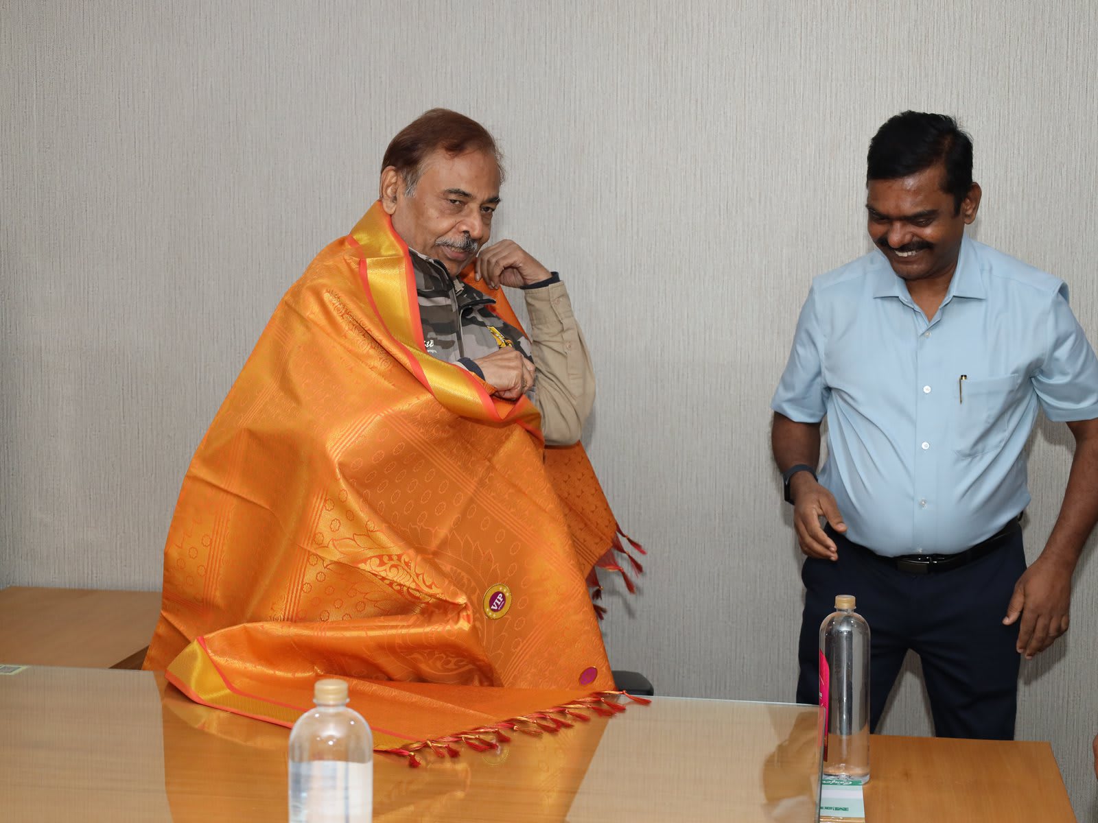 A seated man wrapped in a bright orange silk shawl at a meeting table while another man stands smiling beside him
