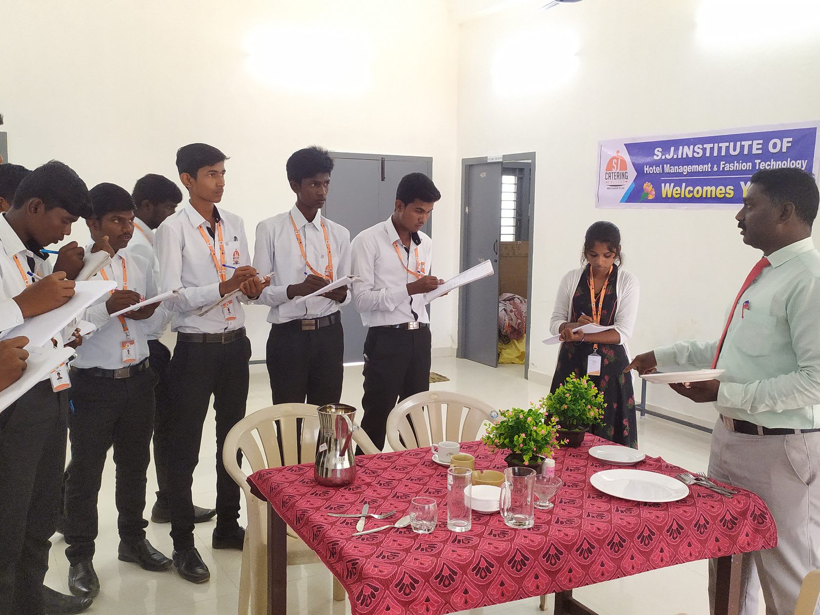 Students in white shirts and ties standing around a small table laid with plates, glasses and cutlery while one pours from a jug and an instructor watches