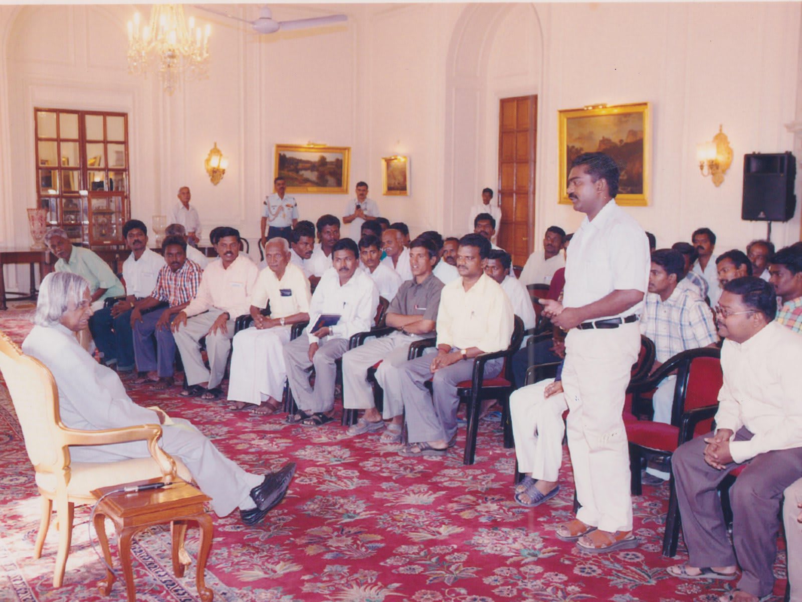 A man in white standing to speak among rows of seated visitors in a chandeliered hall with a patterned red carpet, an elderly man seated in a single chair at the left