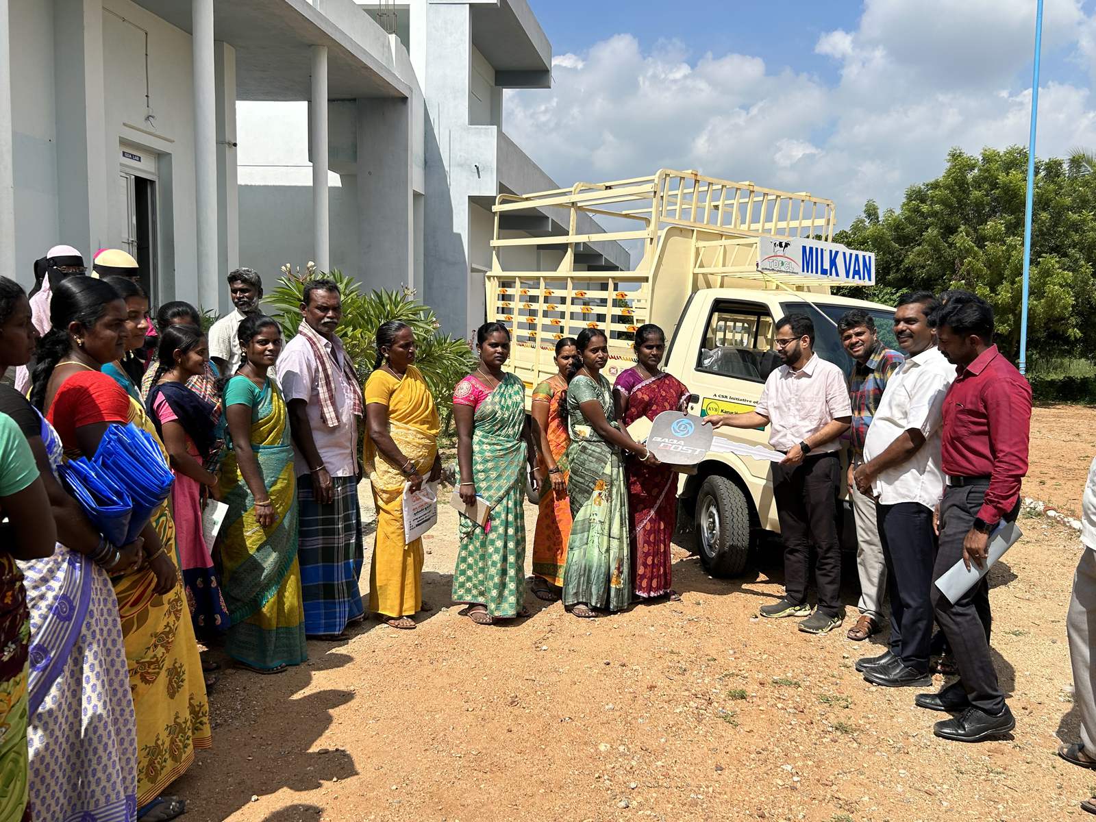 A crowd of women in sarees standing beside a small white pickup marked Milk Van while two men hand over a document