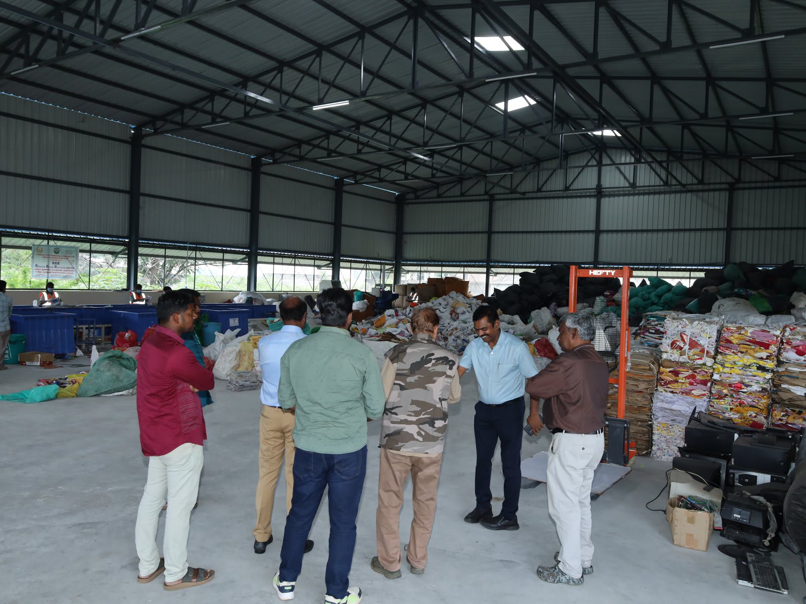 A group of men standing on the concrete floor of a large open shed beside stacked bales of compressed plastic waste and black sacks
