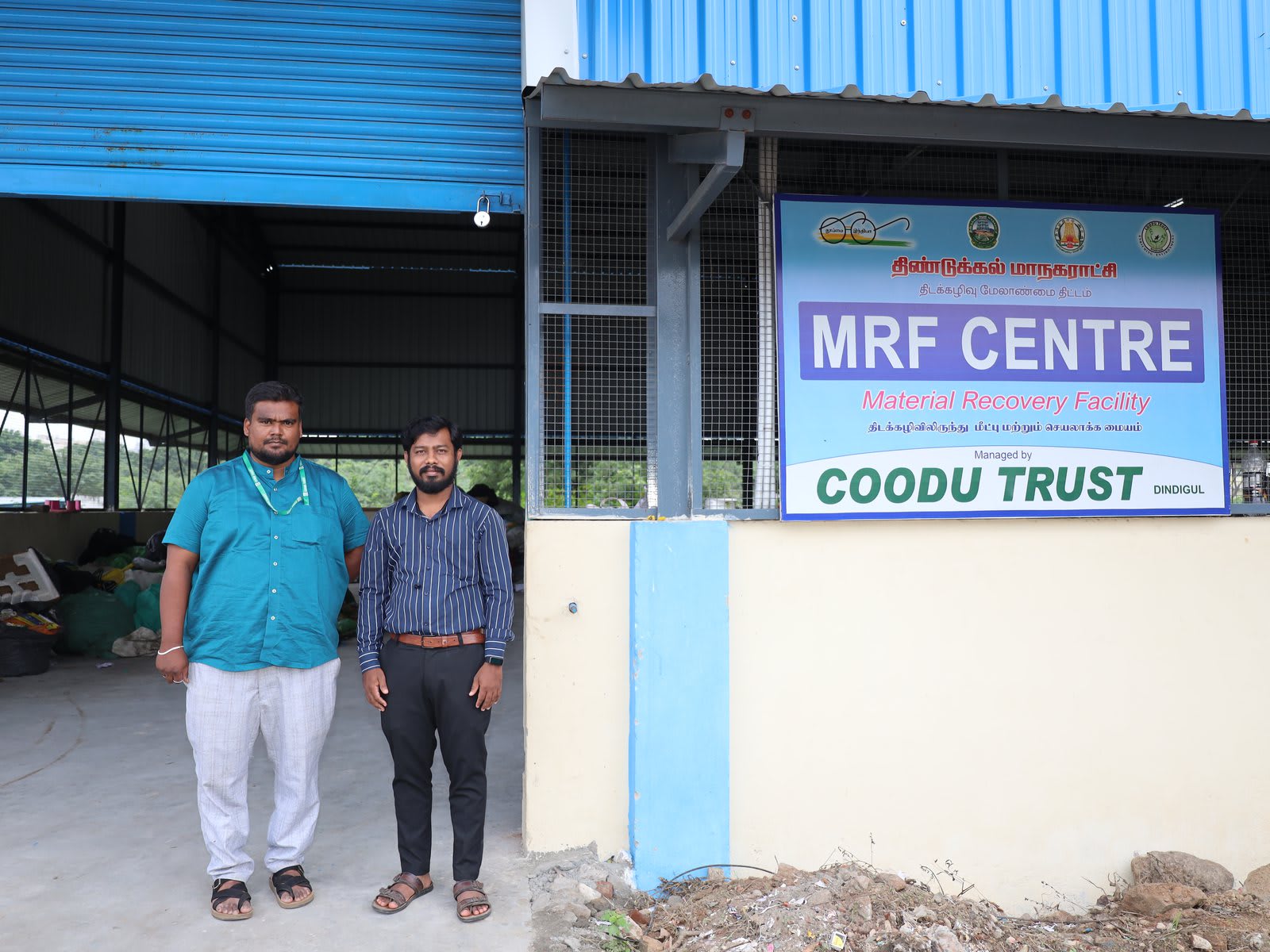 Two men standing in front of a blue and white shed beside a board reading MRF Centre, Material Recovery Facility, managed by Coodu Trust, Dindigul