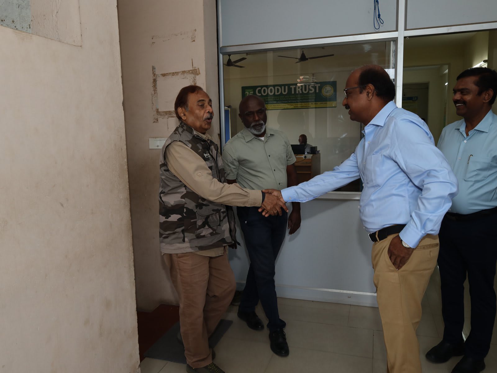 Two men shaking hands in a doorway while two others look on, a green Coodu Trust sign on the wall behind