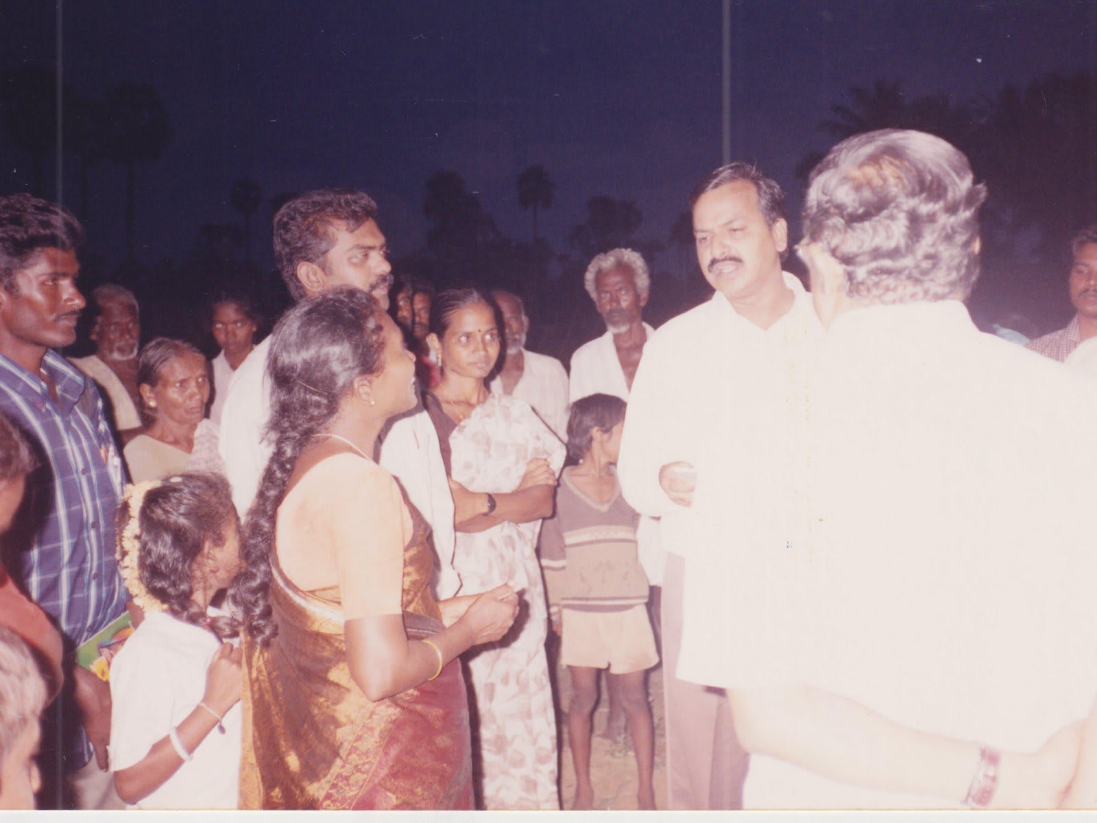 A man in a white shirt standing in a crowd of villagers at dusk, women and children pressed close around him