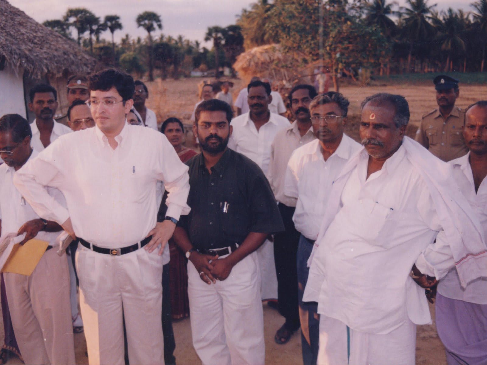 A group of men standing on a bare field beside a thatched hut, one in a white shirt at the front with hands on hips, a police officer at the back