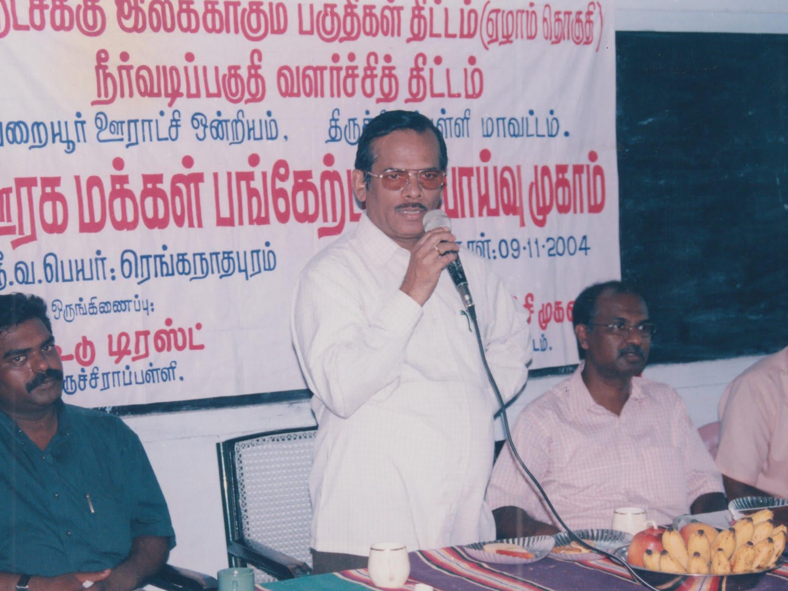 A man speaking into a microphone in front of a large printed Tamil banner, three men seated at a table beside him