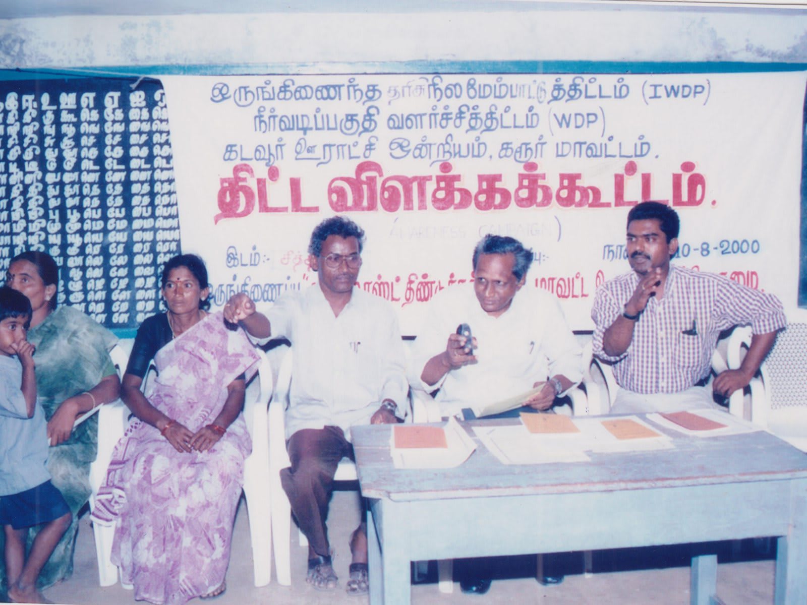Three men and a woman seated at a table under a cloth banner printed in Tamil and English, villagers and children standing at the edges