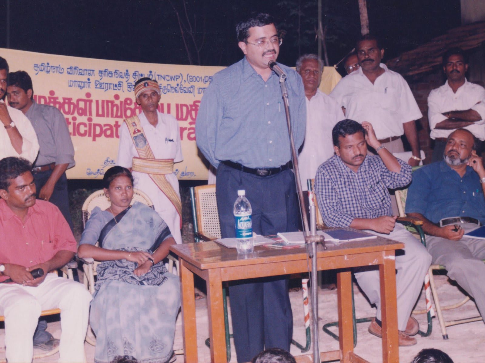 A man in a grey shirt speaking at a microphone beside a table, with villagers seated on chairs and on the ground around him