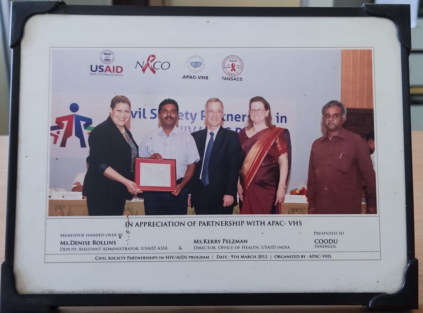 A framed photo-plaque: five people at a memento handover in front of a Civil Society Partnerships in HIV/AIDS backdrop, with a printed caption strip beneath the photograph