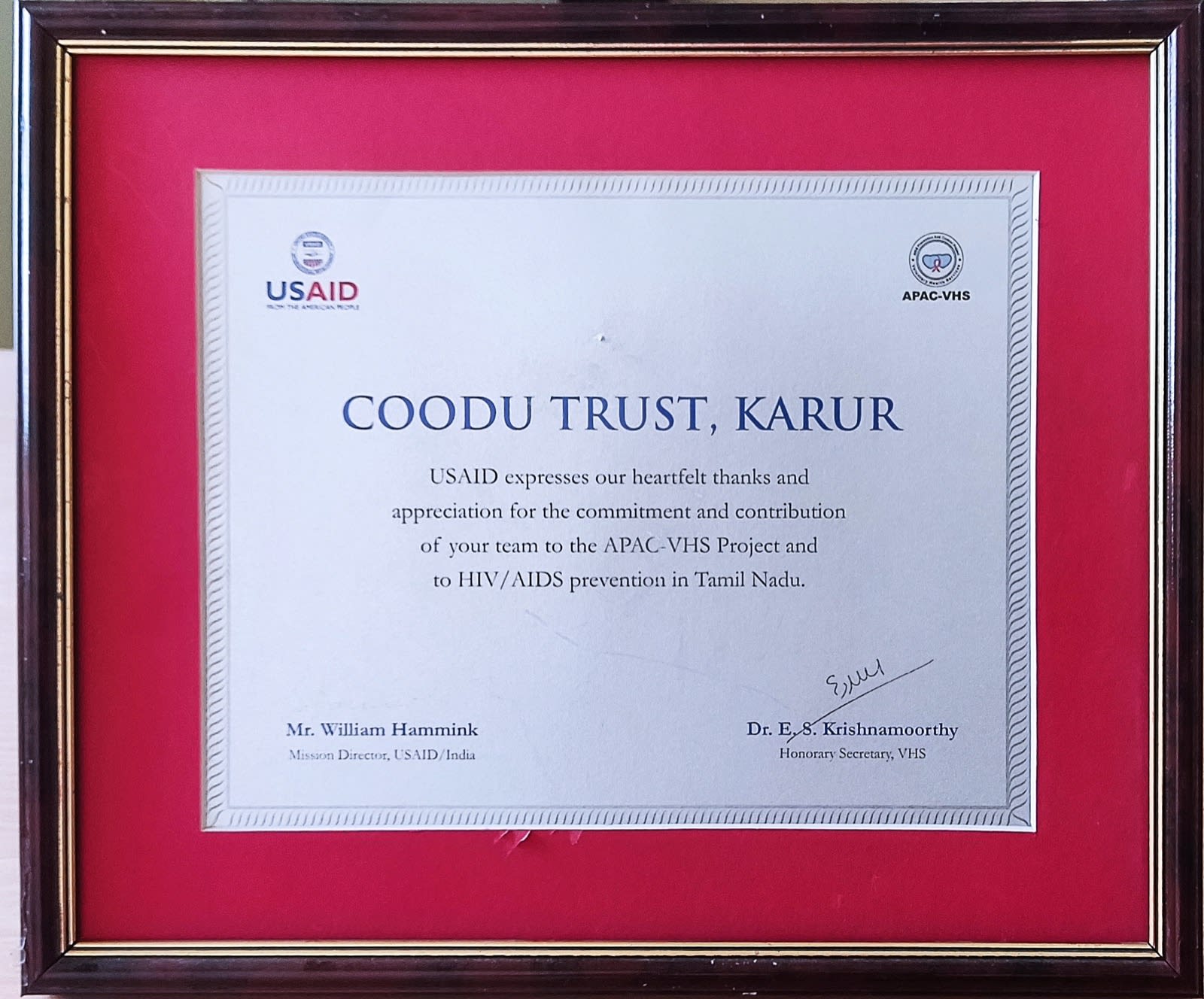A framed certificate on a red mount, headed Coodu Trust, Karur, with the USAID and APAC-VHS logos above the text and two signatures below