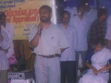 A man speaking into a microphone in front of a banner reading Participatory Rural Appraisal, men standing behind him and a woman seated to the right