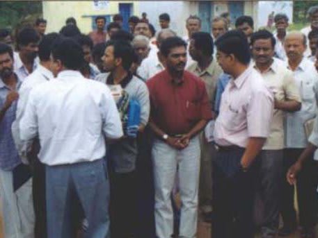 A dense group of men standing outdoors in bright sun, most in white shirts, one at the centre holding papers