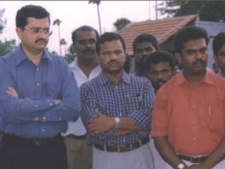 A man in a blue shirt with folded arms standing in front of a group of men outdoors, tiled roofs and trees behind