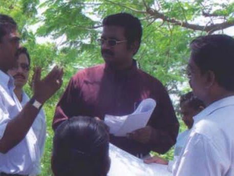 A man gesturing as he speaks to a group of men standing close around him outdoors under trees