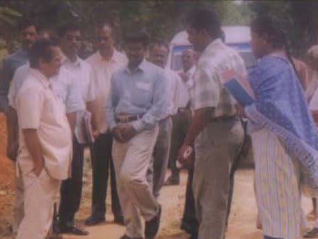Men in light shirts walking across dry scrubland beside a tree, one holding papers