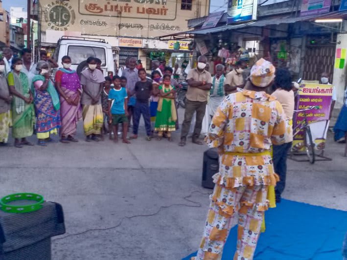 A performer in a patterned costume on a blue mat in a market street, ringed by a standing crowd of shoppers in front of shopfronts