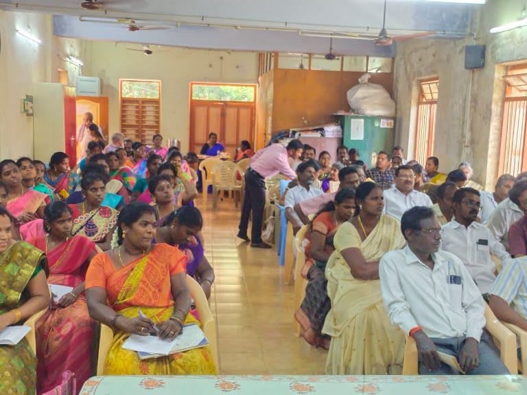 Rows of men and women seated on plastic chairs filling a long hall, many holding printed handouts