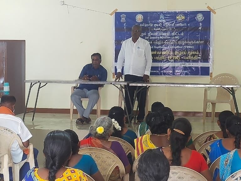 A man standing at a table in front of a blue printed banner addressing about twenty villagers seated on chairs