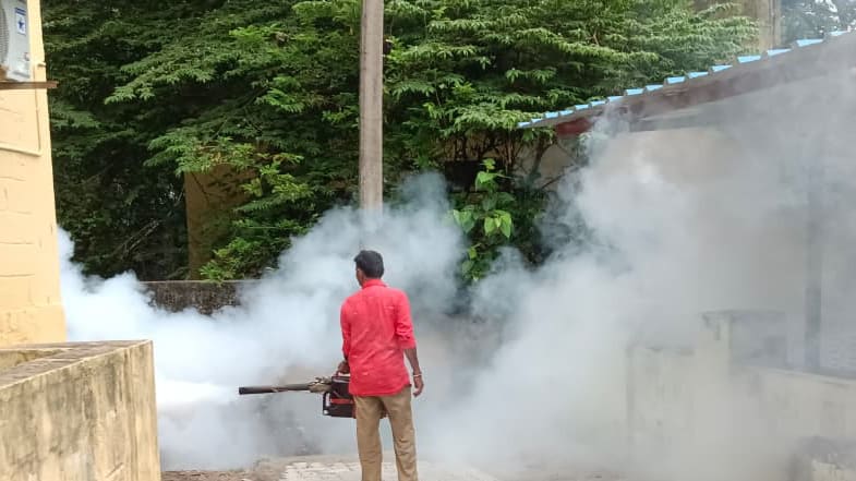 A worker walking a paved lane operating a backpack thermal fogger, dense fog filling the lane