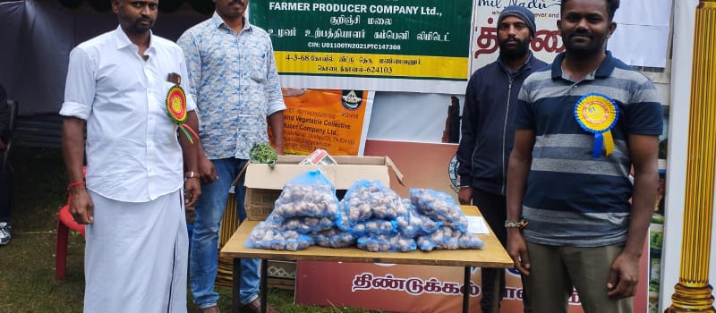 Men stand beside a stall table stacked with mesh bags of garlic under a board naming a farmer producer company