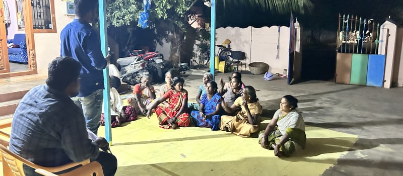 Women seated on a mat in a lit courtyard at night talk with a facilitator standing beside them