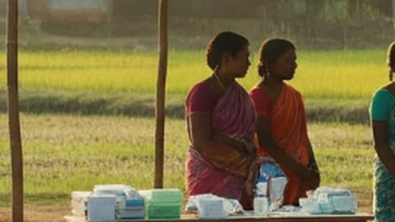 Women waiting at a village health camp set up beside a field, with supplies laid out on a table