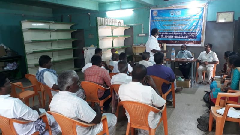 Farmers seated on orange plastic chairs in a produce godown with empty steel shelving, listening to a speaker standing beside a Tamil-language meeting banner, two office bearers at a table to his right