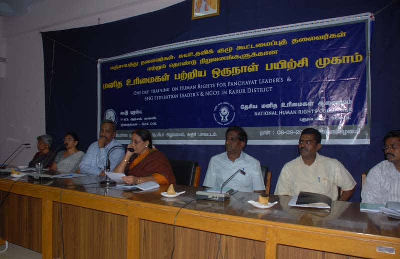A panel of men and women seated at a long table beneath a blue banner reading One Day Training on Human Rights for Panchayat Leaders and SHG Federation Leaders and NGOs in Karur District