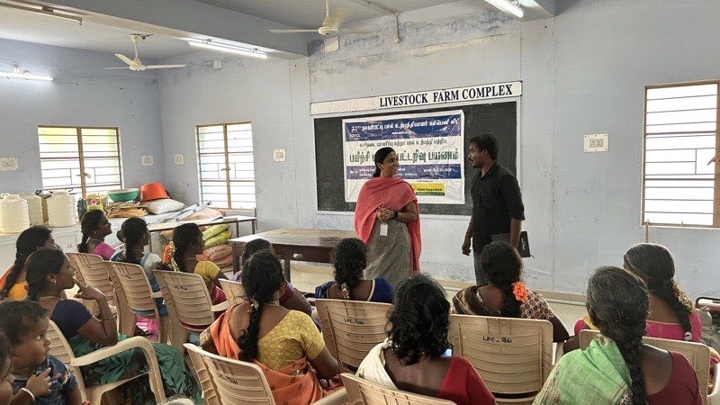 A woman and a man standing in front of a banner, speaking to a room of women seated in plastic chairs