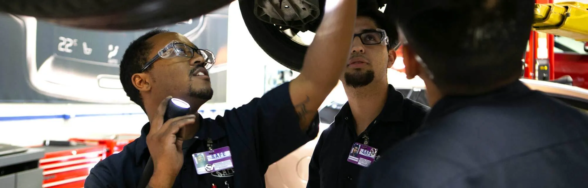 Three auto technician or mechanic students from the Volvo program at Universal Technical Institute Avondale who are looking at a Volvo that is up on a lift