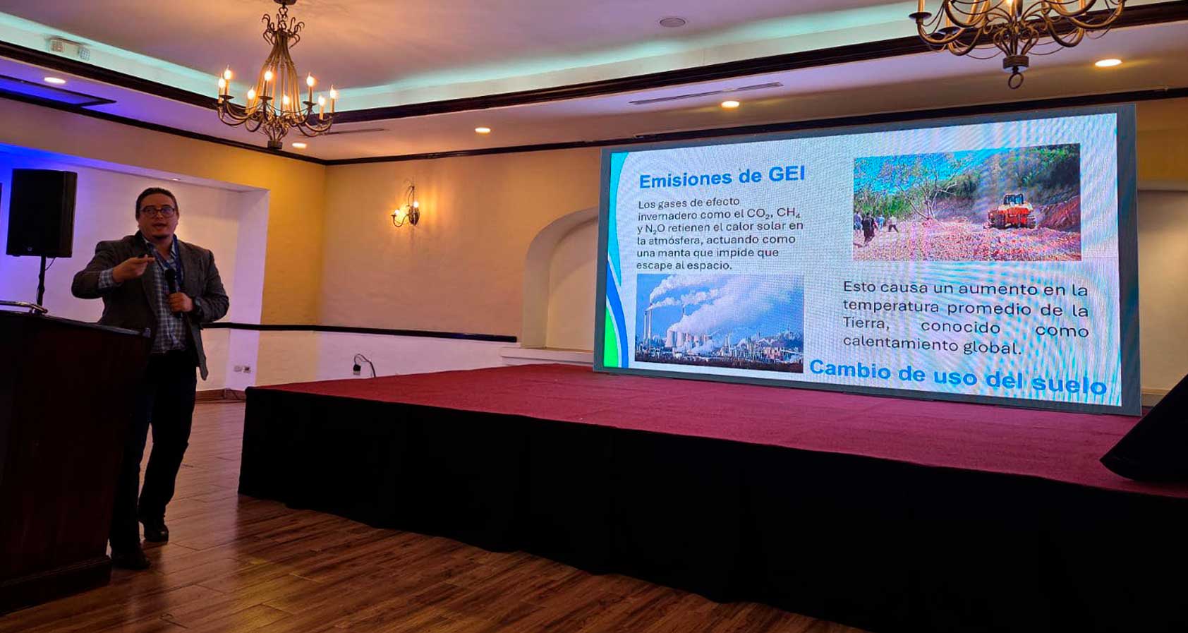 Hombre con micrófono ofrece una conferencia sobre emisiones de gases de efecto invernadero en un salón; al fondo se proyecta una diapositiva que explica cómo el CO₂, CH₄ y N₂O retienen el calor en la atmósfera, contribuyendo al calentamiento global y al cambio de uso del suelo.