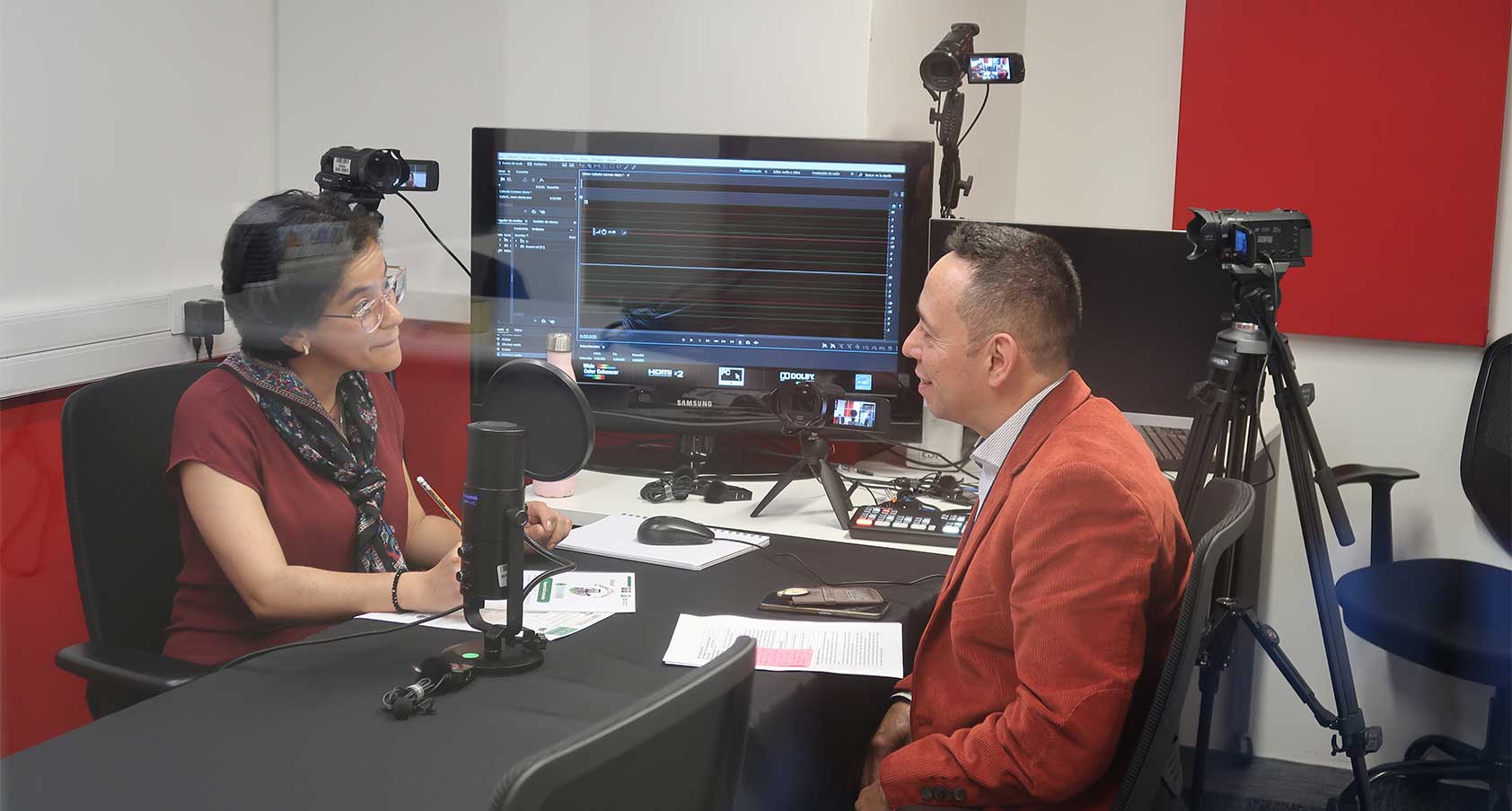 “En un estudio de grabación, una mujer con blusa oscura y bufanda platica con un hombre de saco naranja sentado frente a ella. Entre ambos hay un micrófono de podcast sobre la mesa, con hojas y cables, mientras detrás se observan cámaras, un control de audio y una pantalla con una línea de tiempo de edición.”