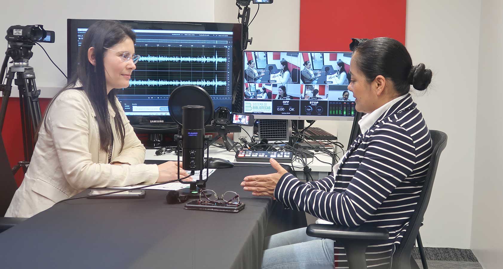 “Dos personas sentadas frente a frente en una mesa, en la que tienen un micrófono de podcast. La mujer a la izquierda viste chaqueta clara y gafas, mientras que la persona a la derecha —con cabello recogido y suéter de rayas— gesticula con las manos. Al fondo, se aprecia un monitor con ondas de audio y equipo de grabación.”