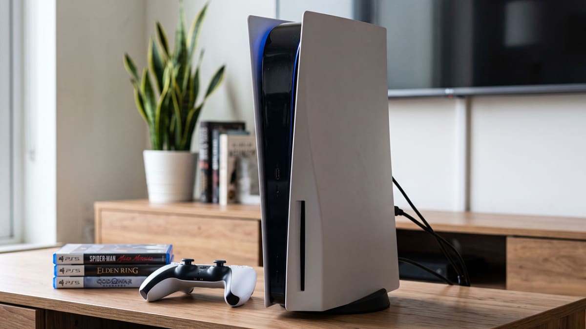 A disc-edition PS5 console on a wooden TV unit beside a stack of physical PS5 game discs and a DualSense controller