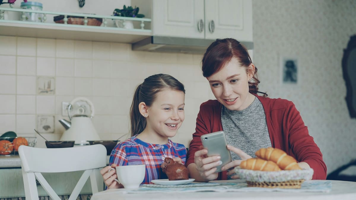 Mother and young daughter looking at a phone together at the kitchen table, preparing for the NZ social media ban