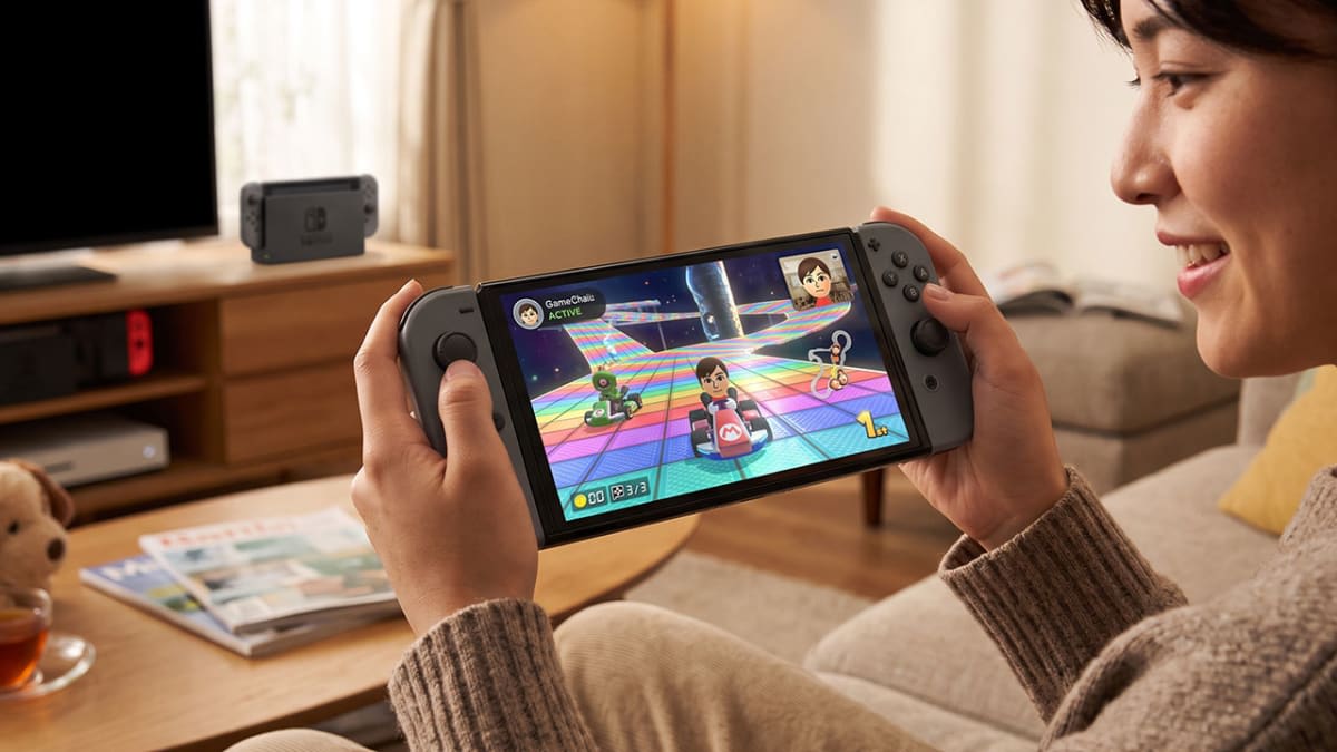 Person playing a Nintendo Switch 2 in handheld mode on a couch, with the dock and TV in the background