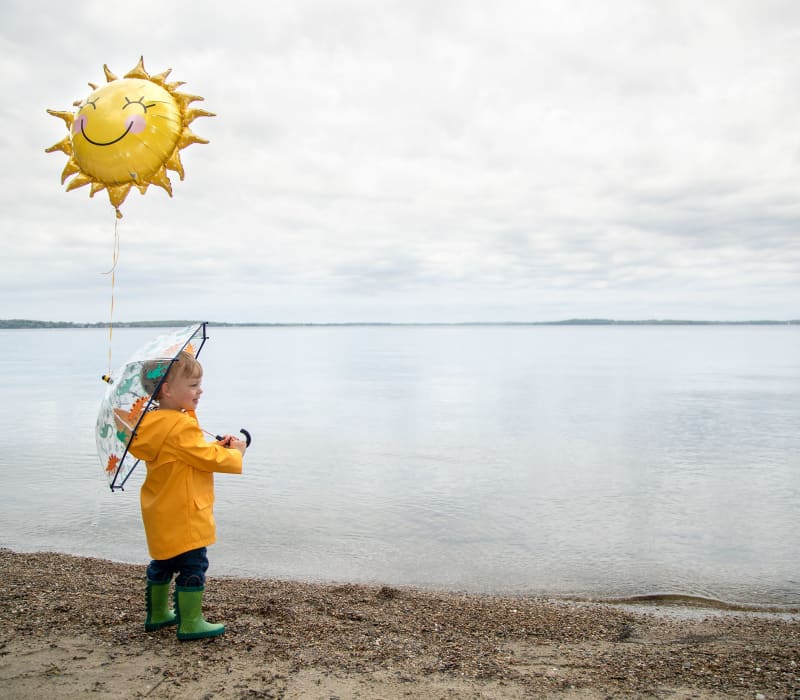 Rykal Wasson 343273-1022 Small boy on the shore of a lake, dressed in a raincoat and holding a balloon.
