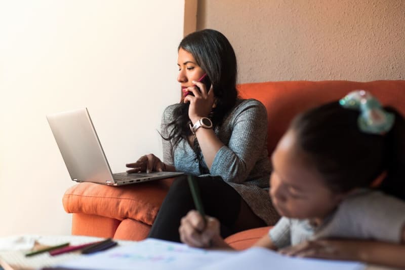 Individual sitting on sofa with their laptop and talking on phone while child does homework in foreground