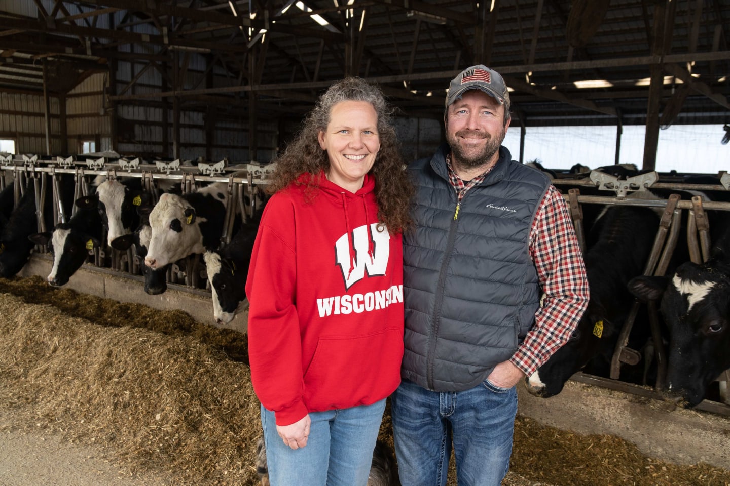 Woman in Wisconsin Badgers sweatshirt next to man in jacket, both smiling and standing in front of cows feeding in a barn.