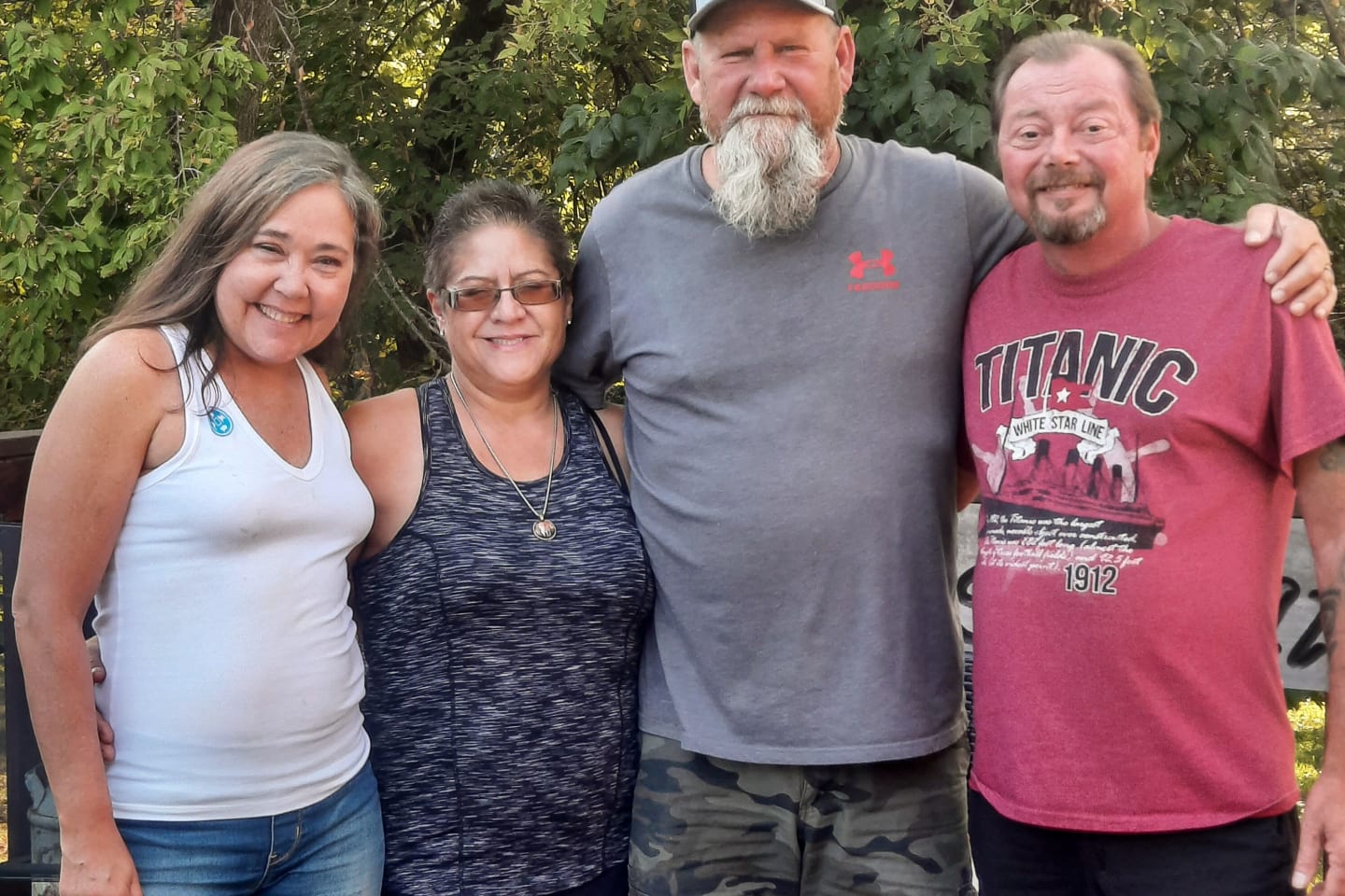 Four adults, two women and two men, facing the camera smiling with their arms around each other.